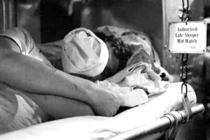 1957-Operation-Strike-Back_300_200 A young boy in a scouting uniform lies on a narrow bunk bed inside what appears to be a ship or submarine, smiling at the camera.