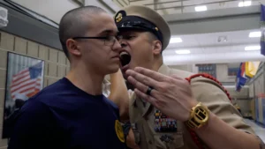 A military officer in uniform yells at a young recruit, who stands still and shows no emotion, inside a building with flags and photos in the background.