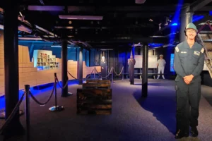A dimly lit museum exhibit with mannequins dressed in naval uniforms, ropes sectioning off areas, wooden crates, and blue lighting creating a dramatic atmosphere.