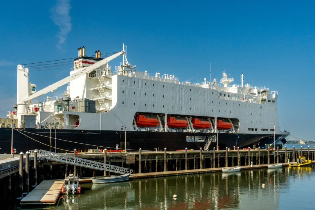 A large white ship with red lifeboats, often used for training by Maritime Academies and schools, is docked at a pier under a clear blue sky, with gangways connecting it to the shore and boats in the water nearby.