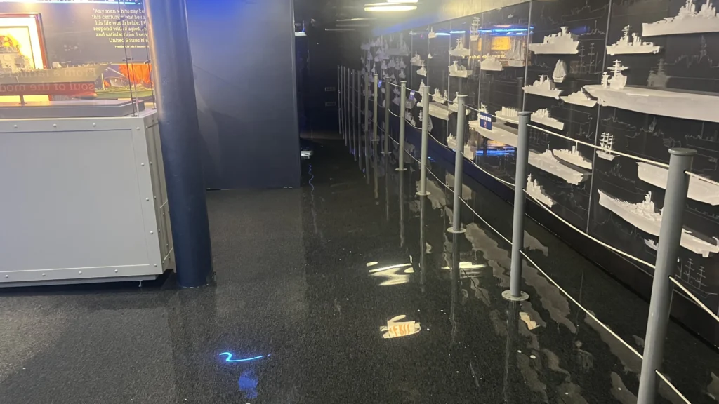 A hallway with a shiny black floor reflects lights above. On the right, rows of detailed ship models are displayed on shelves behind barriers. The hallway appears empty, and an exit sign is visible near the floor.