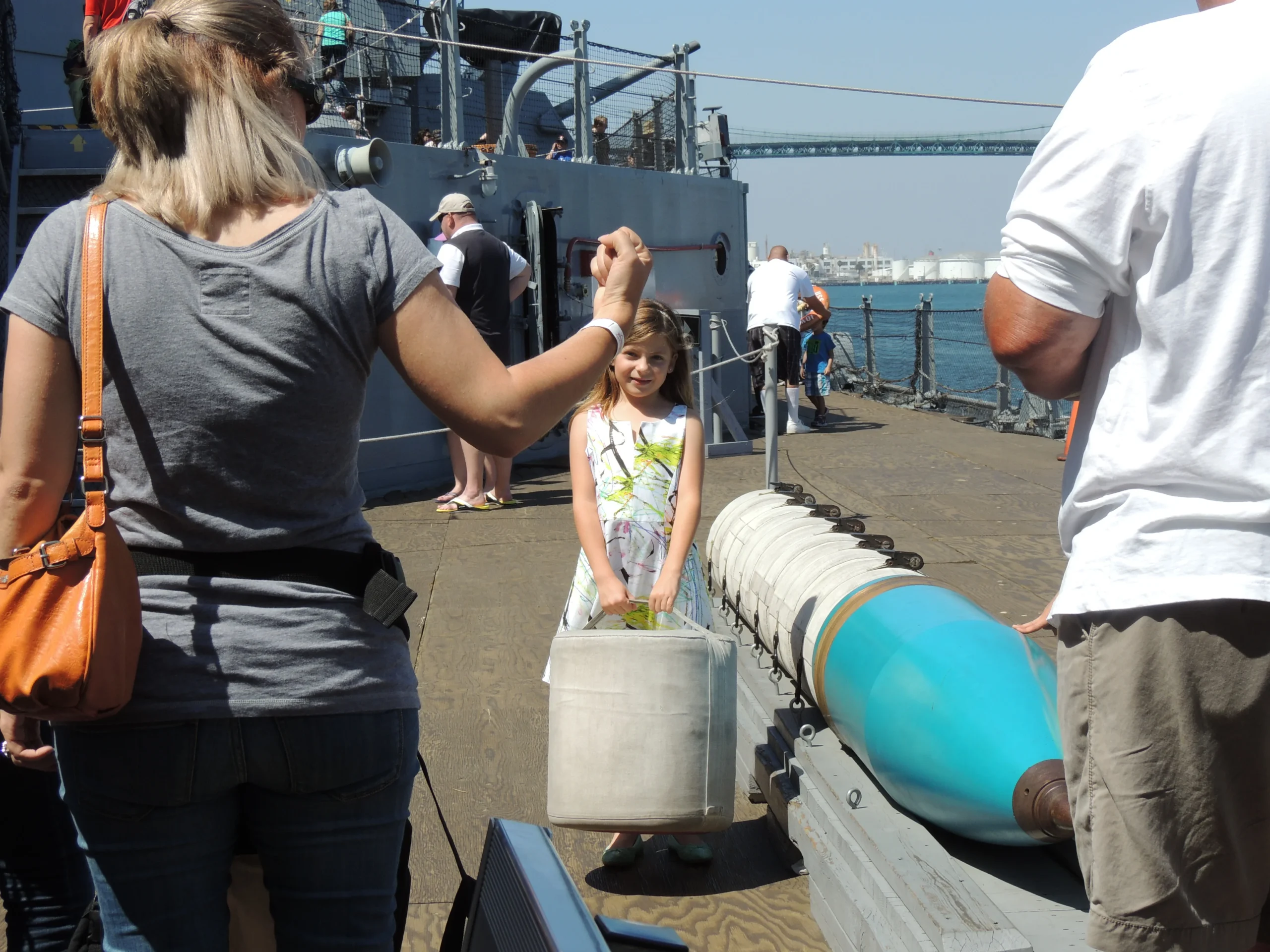 A young girl in a sundress stands on a ships deck, smiling, with adults nearby. A large blue and white torpedo is displayed beside her. A bridge and waterfront are visible in the background. A young girl in a sundress stands on a ships deck, smiling, with adults nearby. A large blue and white torpedo is displayed beside her. A bridge and waterfront are visible in the background.