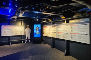 A person in a naval uniform stands in a dimly lit room with informational panels on the walls and a digital display at the center. Blue lighting highlights the industrial ceiling pipes.
